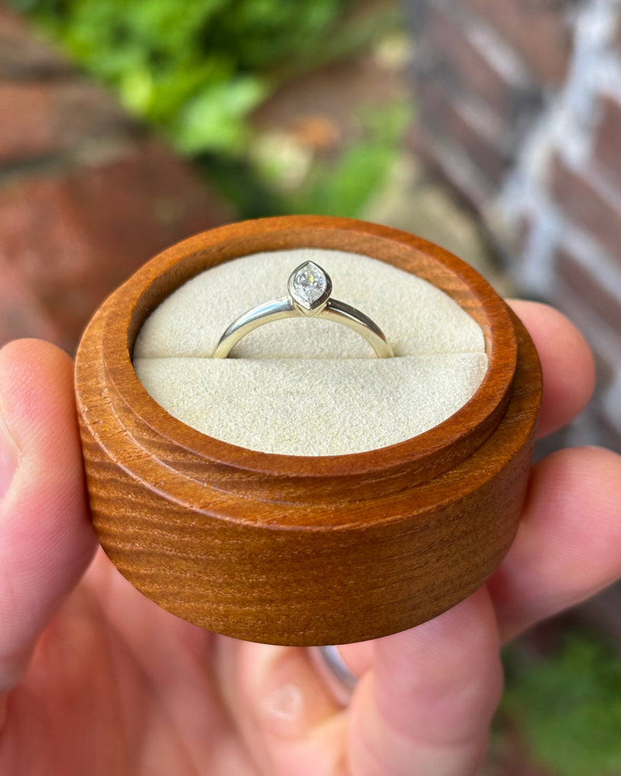 Marquise cut diamond engagement ring in a silver bezel setting, held in a round wooden box.