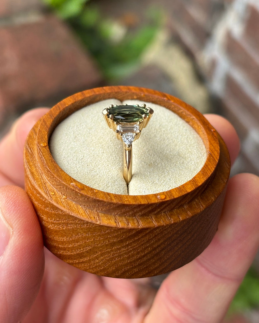 Marquise cut green sapphire engagement ring with diamond side stones set in a gold band in a wooden box.