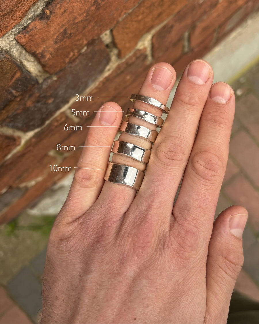 Silver wedding bands in varying widths from 3mm to 10mm worn on a hand against a brick background.