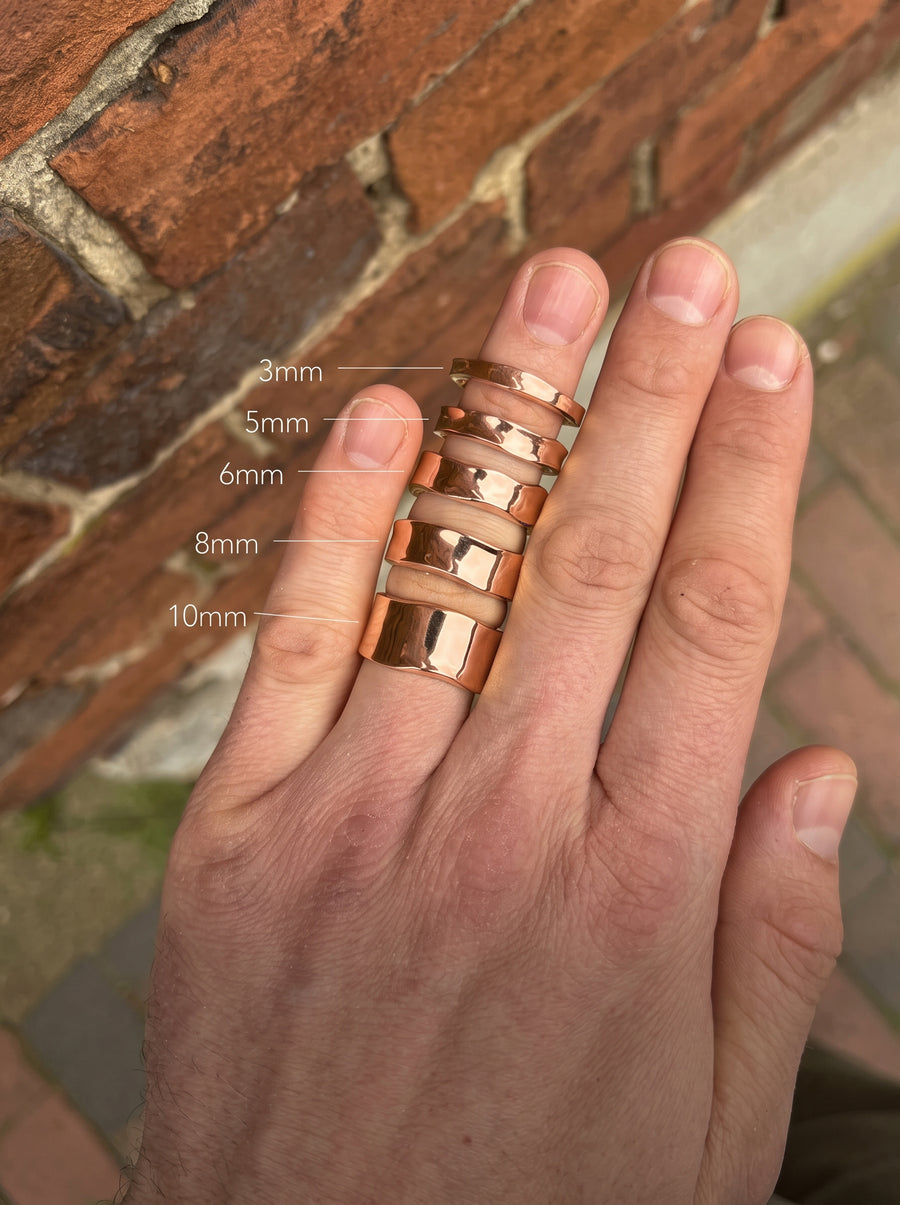 Rose gold wedding rings in various widths from 3mm to 10mm worn on a hand against a brick background.