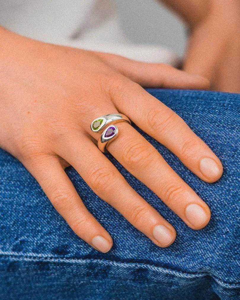 Hand wearing a silver ring with peridot and amethyst gemstones on blue jeans