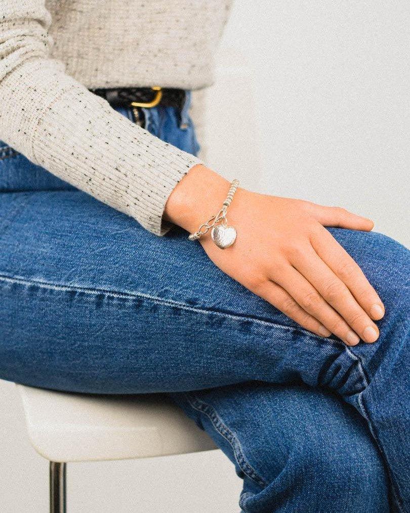 Hammered silver heart charm bracelet on wrist, paired with casual jeans and a sweater