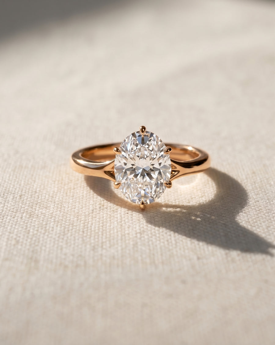 Oval cut diamond engagement ring in 18ct rose gold with a tulip claw setting on a textured fabric background.