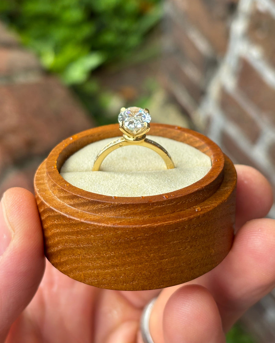 Oval lab-grown diamond solitaire engagement ring with a gold pavé band in a wooden ring box.