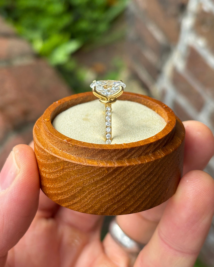 Oval lab-grown diamond solitaire ring with a pavé band set in yellow gold, held in a wooden box.