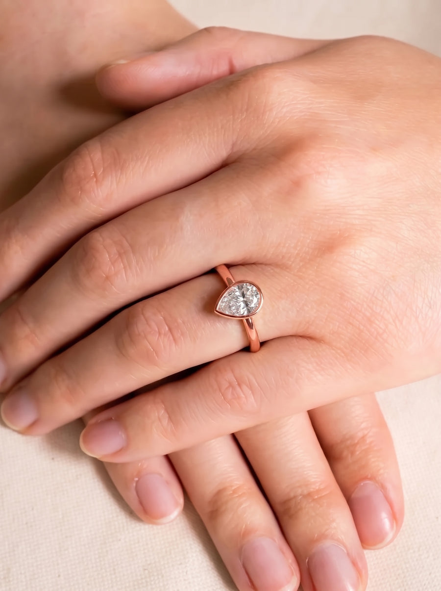 Pear cut diamond engagement ring in a rose gold bezel setting worn on a finger.