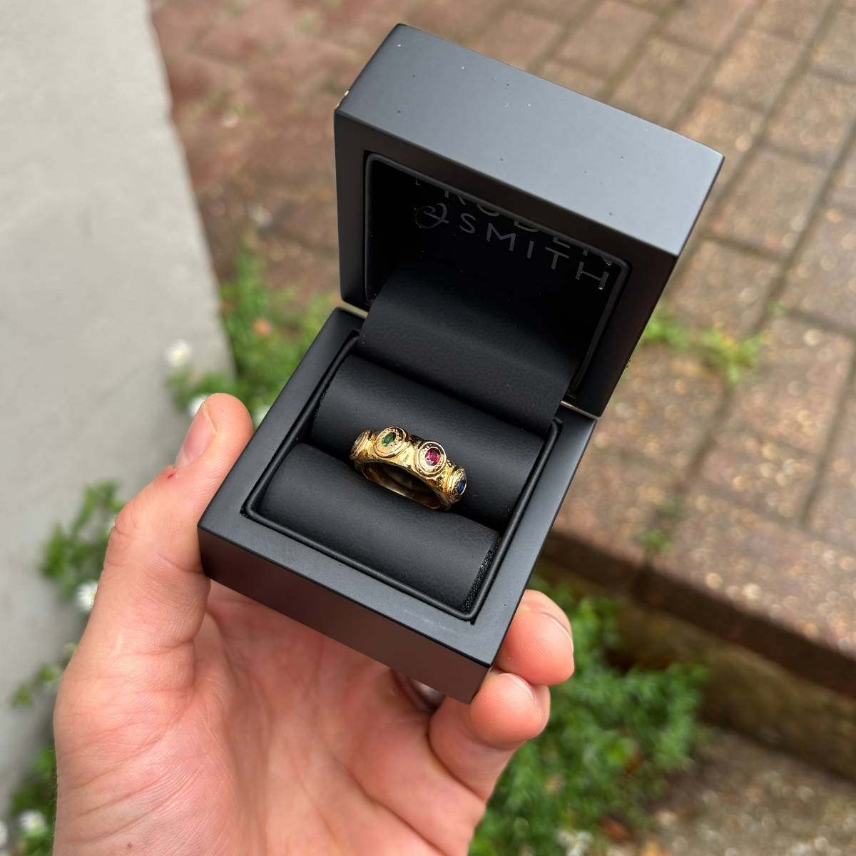 A gold ring set with multiple coloured gemstones, presented in a black Pruden and Smith jewellery box and held in a hand.