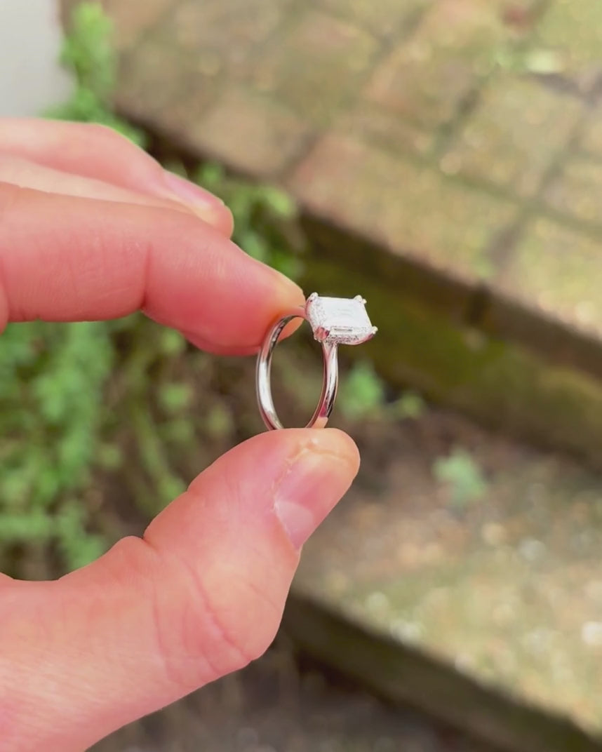 emerald cut diamond hidden halo engagement ring behind rotated with green leafy background