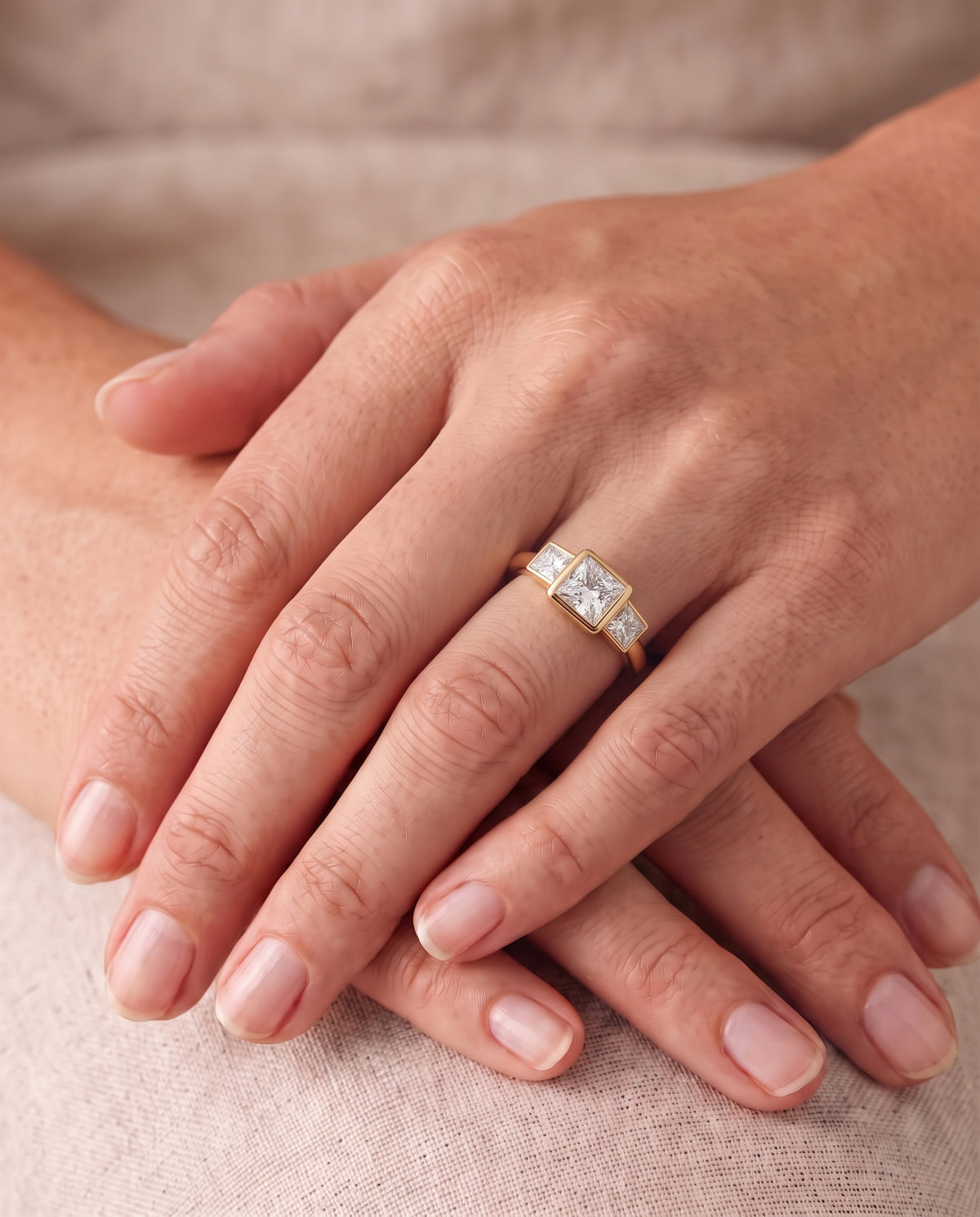 Gold trilogy engagement ring with three princess cut diamonds in a bezel setting on a finger.