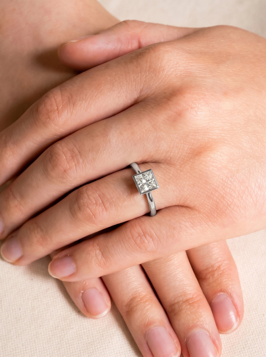 Princess cut diamond engagement ring in a silver-toned bezel setting worn on a finger.