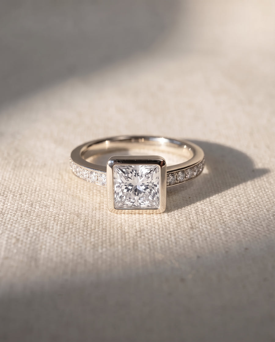 9ct white gold engagement ring with a princess cut center stone in a bezel setting and pavé diamond band.