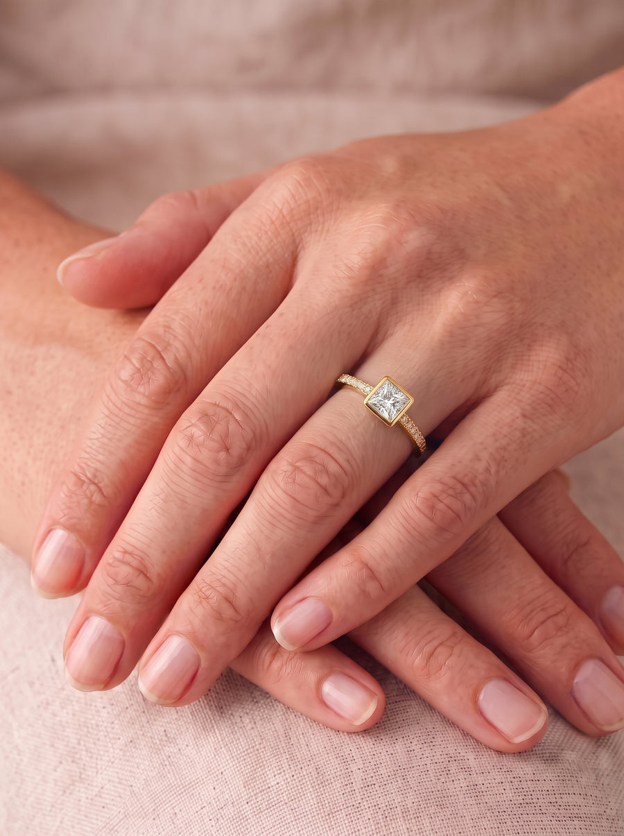 Princess cut diamond engagement ring in a gold bezel setting with a pavé band on a finger.