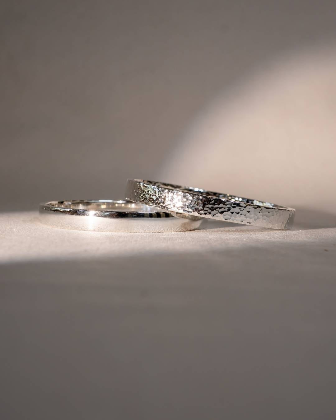 Two handmade silver wedding rings with hammered and polished textures on a neutral background