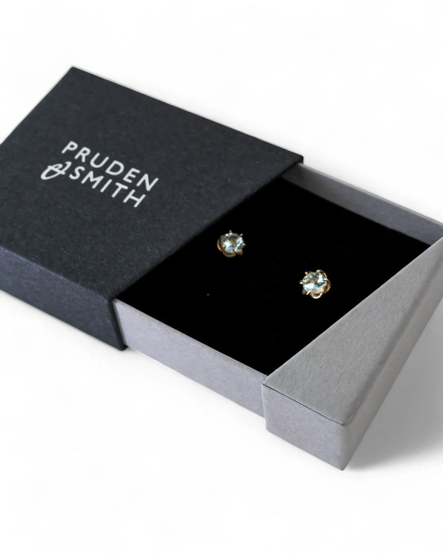 Gold revolved claw stud earrings with light blue gemstones in a black box.