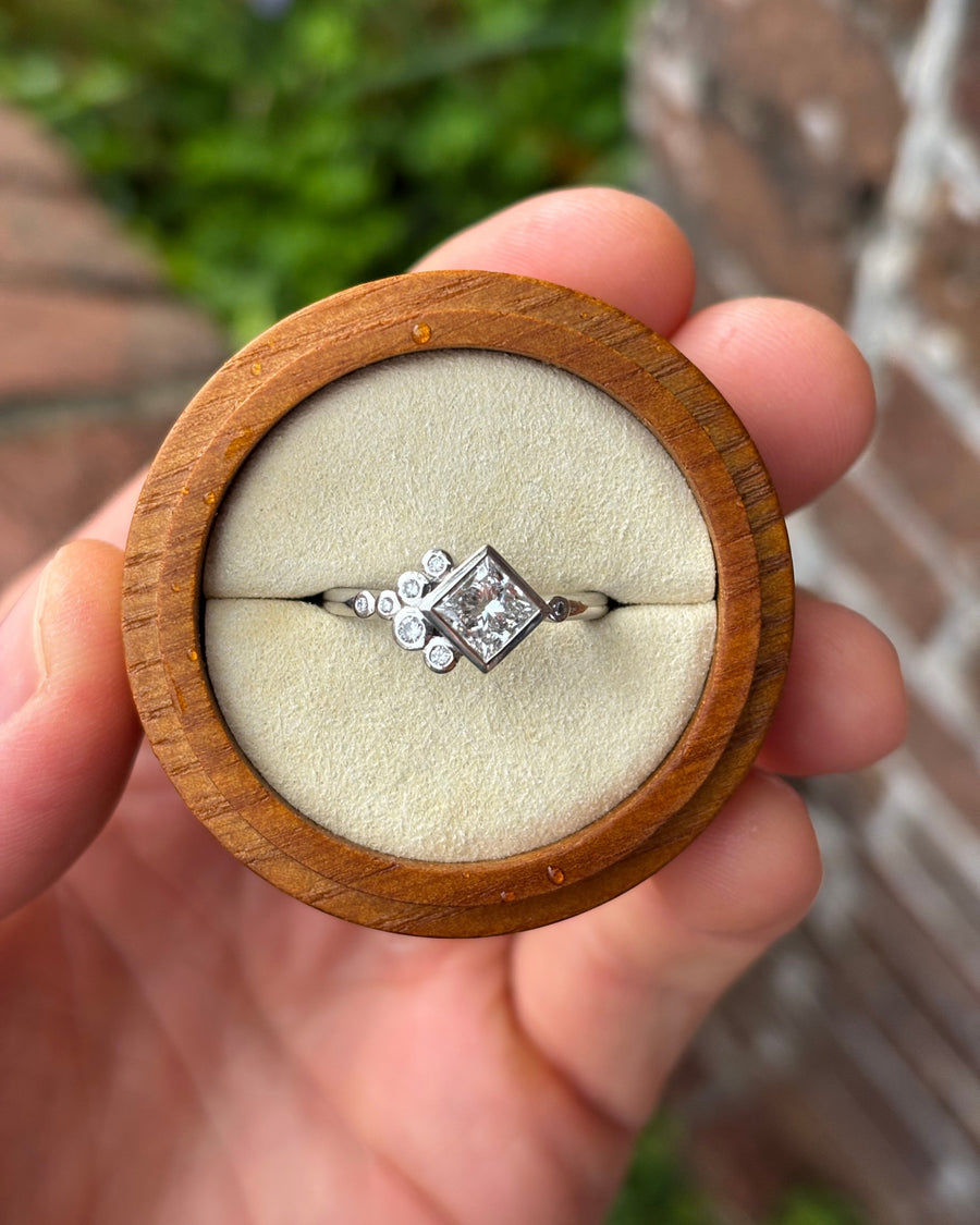 Platinum engagement ring with a square emerald cut diamond and bubble-style accent stones in a wooden box.