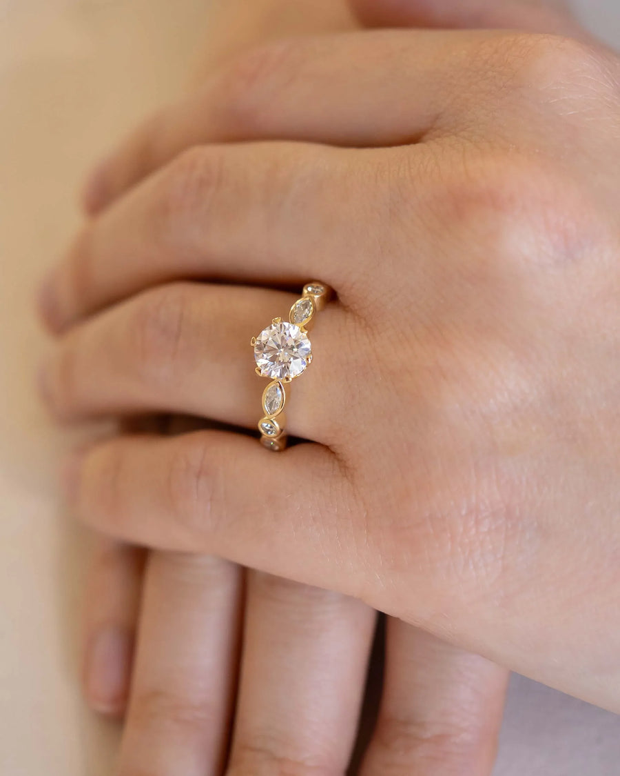 Gold engagement ring with a round cut diamond and marquise side stones on a hand.