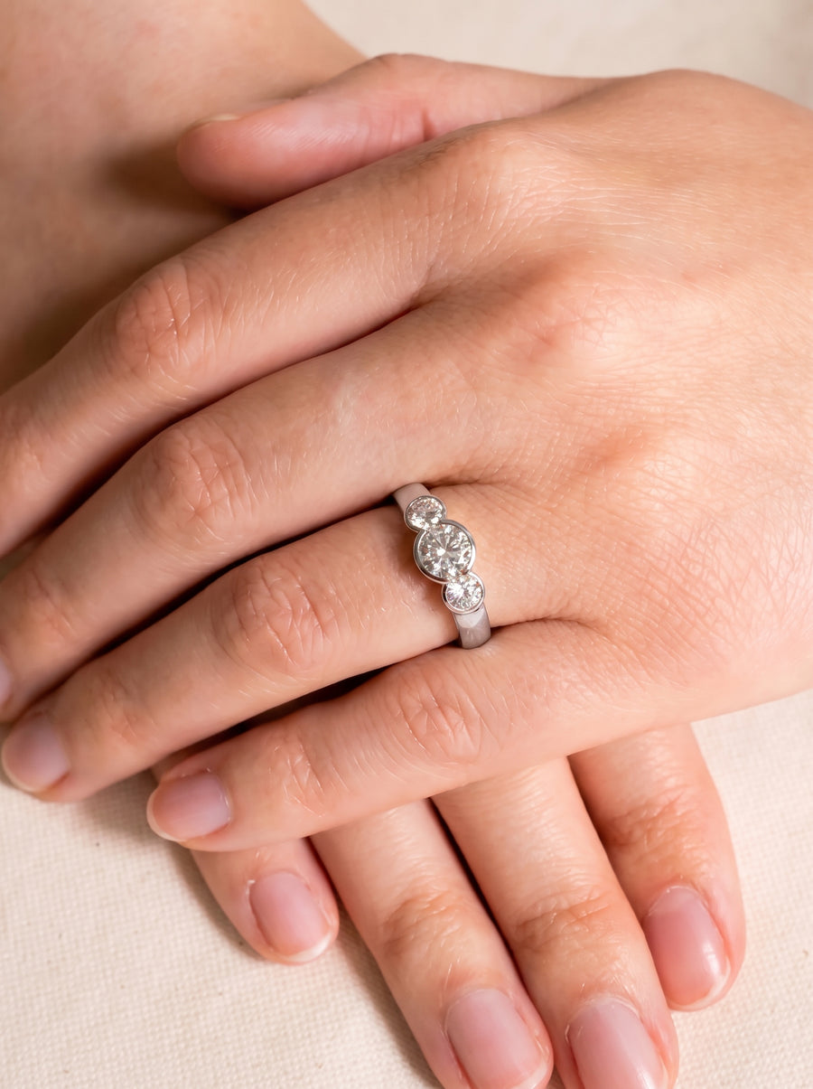 Trilogy engagement ring with three round cut diamonds in a silver-toned bezel setting on a finger.