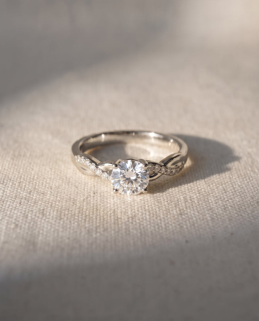 Round cut diamond engagement ring with a silver twisted band and pavé accents on a textured fabric background.