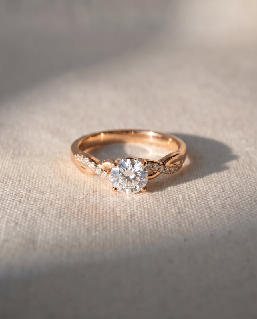 Rose gold engagement ring with a round cut center diamond and a twisted band featuring small pavé diamonds.