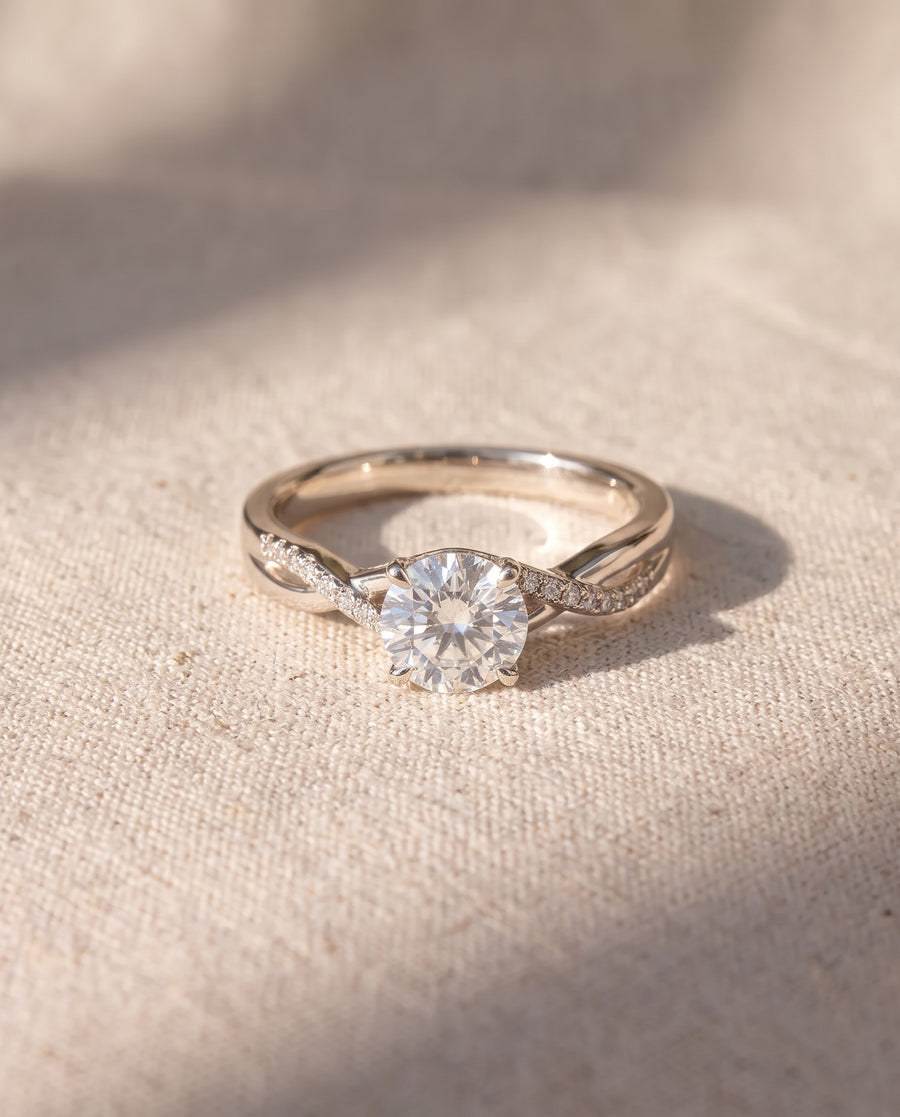 Round cut diamond engagement ring with a twisted silver band featuring pavé accents on a textured fabric background.