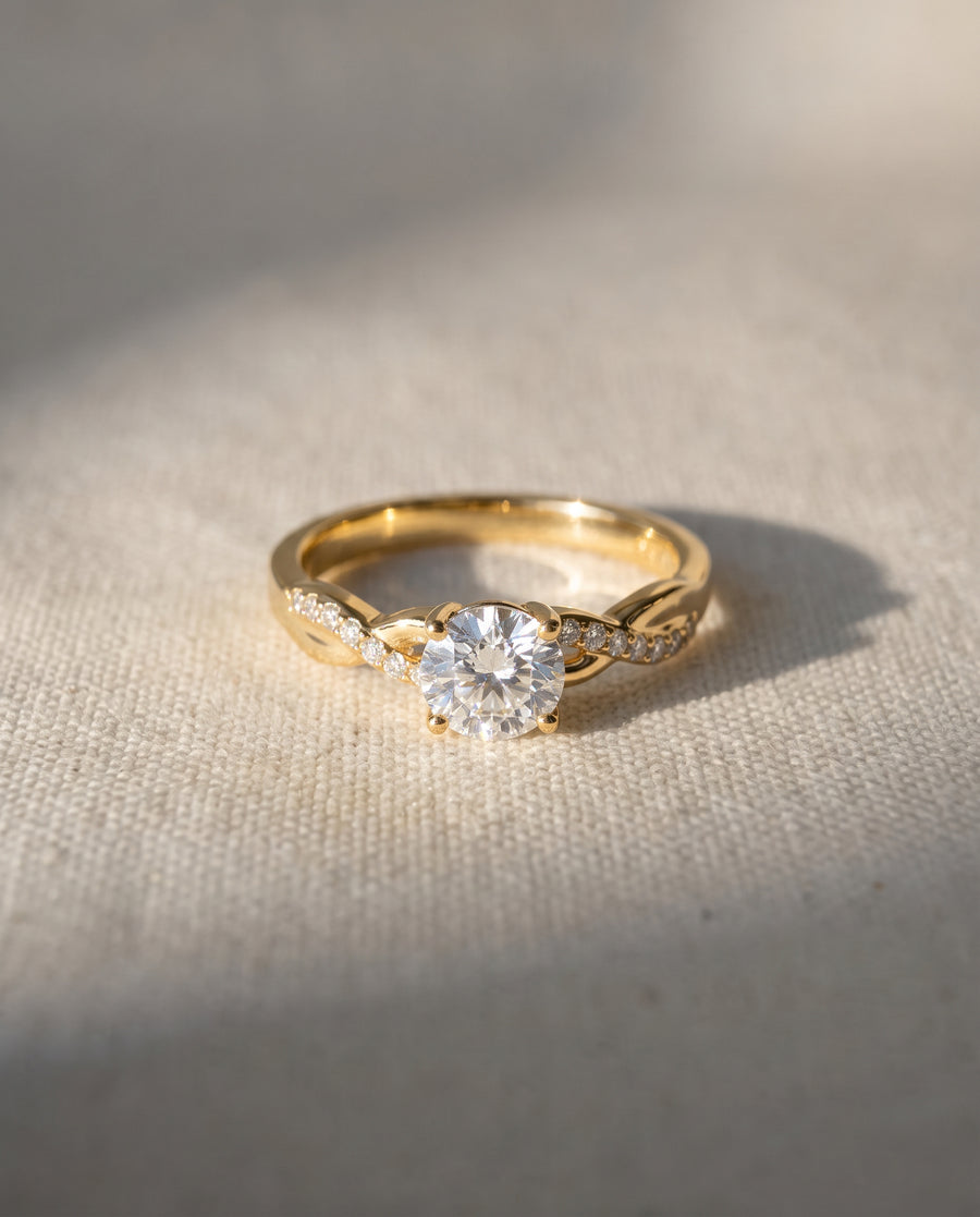 Round cut diamond engagement ring with a gold twisted band and pavé accents on a textured fabric background.