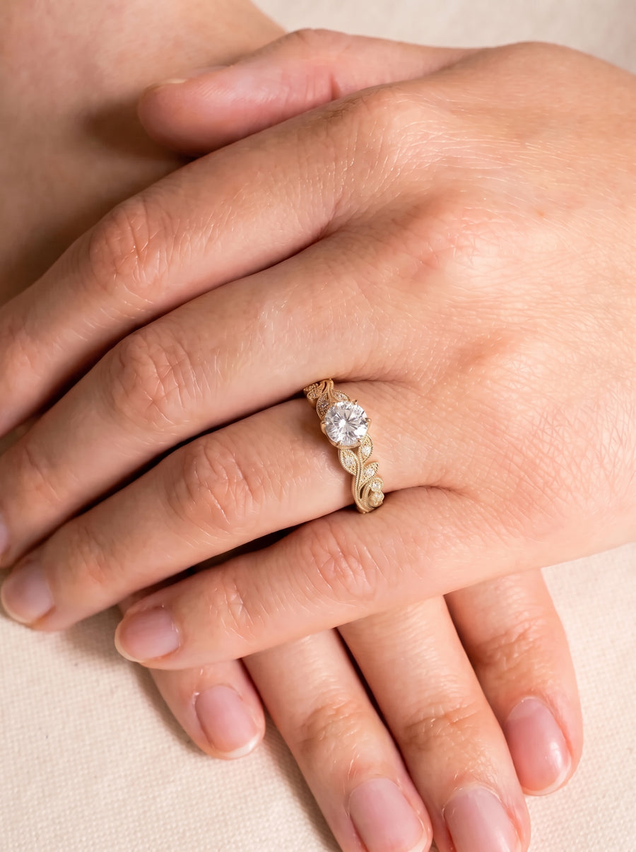 Round cut diamond engagement ring in a gold band with intricate leaf-inspired filigree details on a finger.