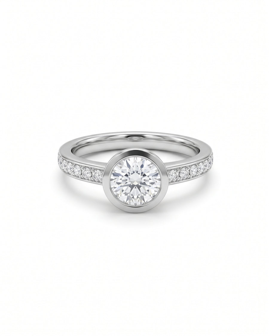 Round diamond engagement ring in a silver-toned metal with a bezel setting and pavé diamond band.