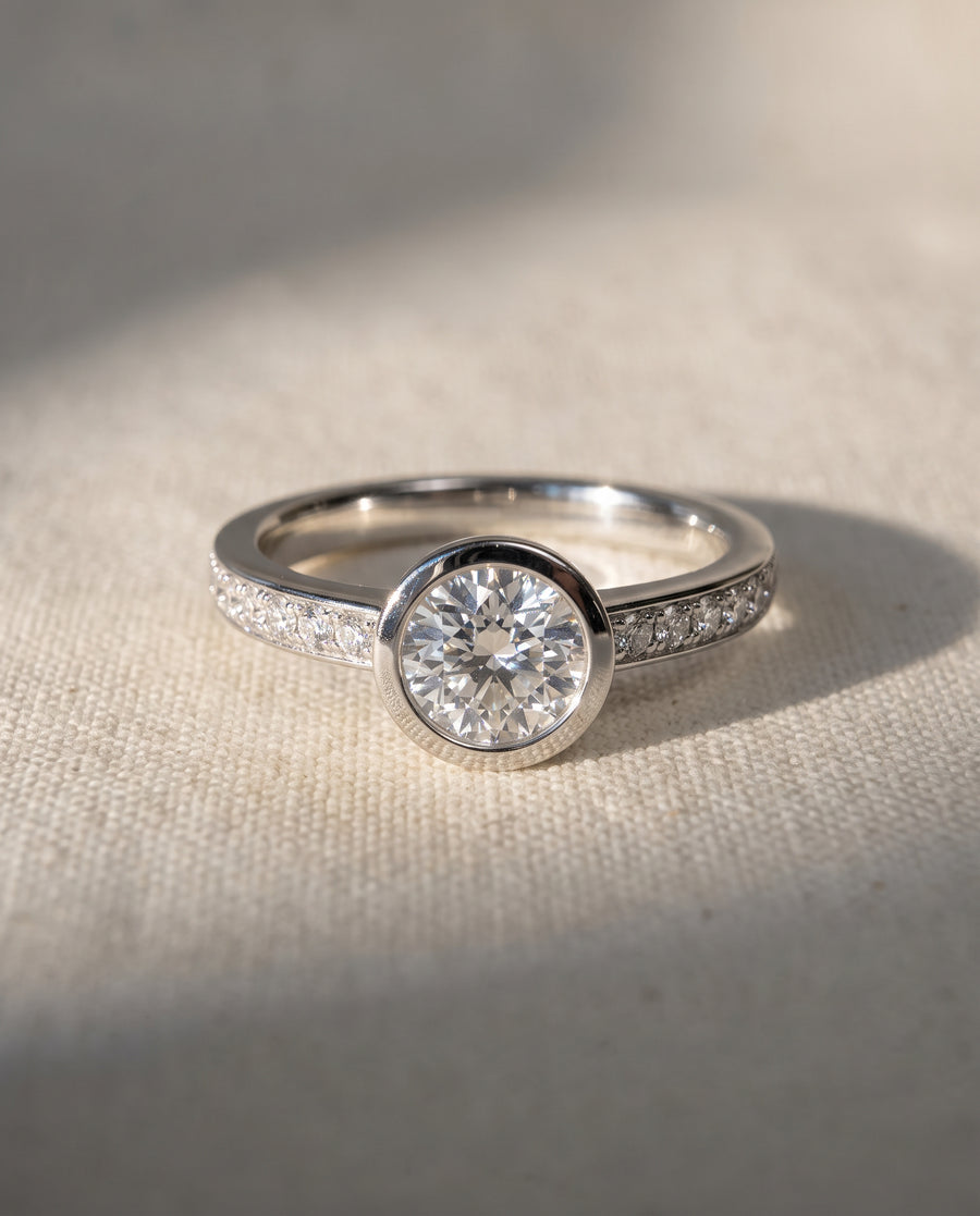 Round diamond engagement ring in 18ct rose gold with a bezel setting and pavé diamond band.