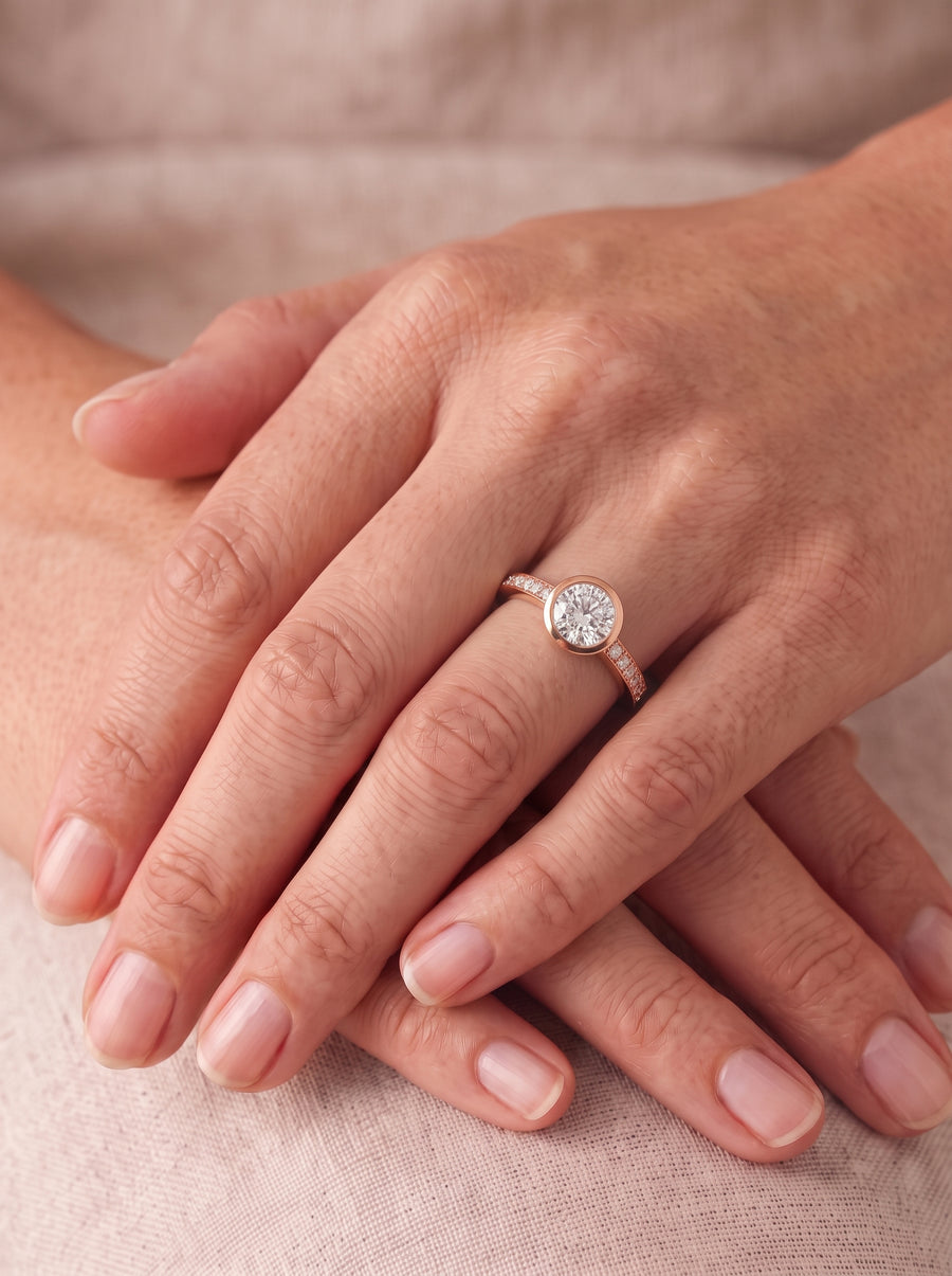 Round diamond engagement ring in a rose gold bezel setting with a pavé band on a finger.