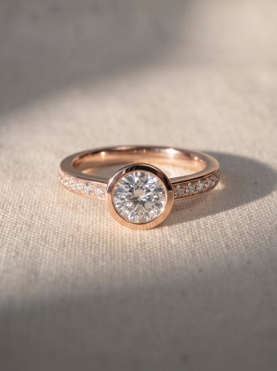 Round diamond engagement ring in 9ct rose gold with a bezel setting and pavé diamond band.