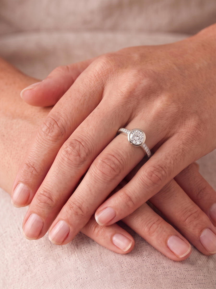 Round diamond engagement ring with a bezel setting and pavé band in white gold on a finger.