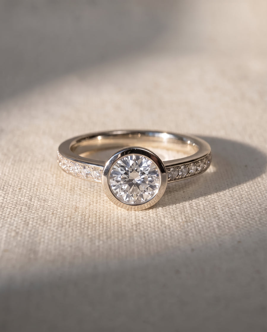 Round diamond engagement ring in 9ct white gold with a bezel setting and pavé diamond band.
