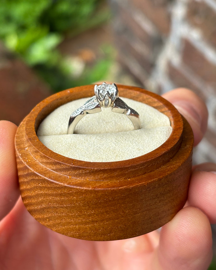 Round cut diamond engagement ring in a silver-toned band with vintage leaf-style detailing in a wooden box.
