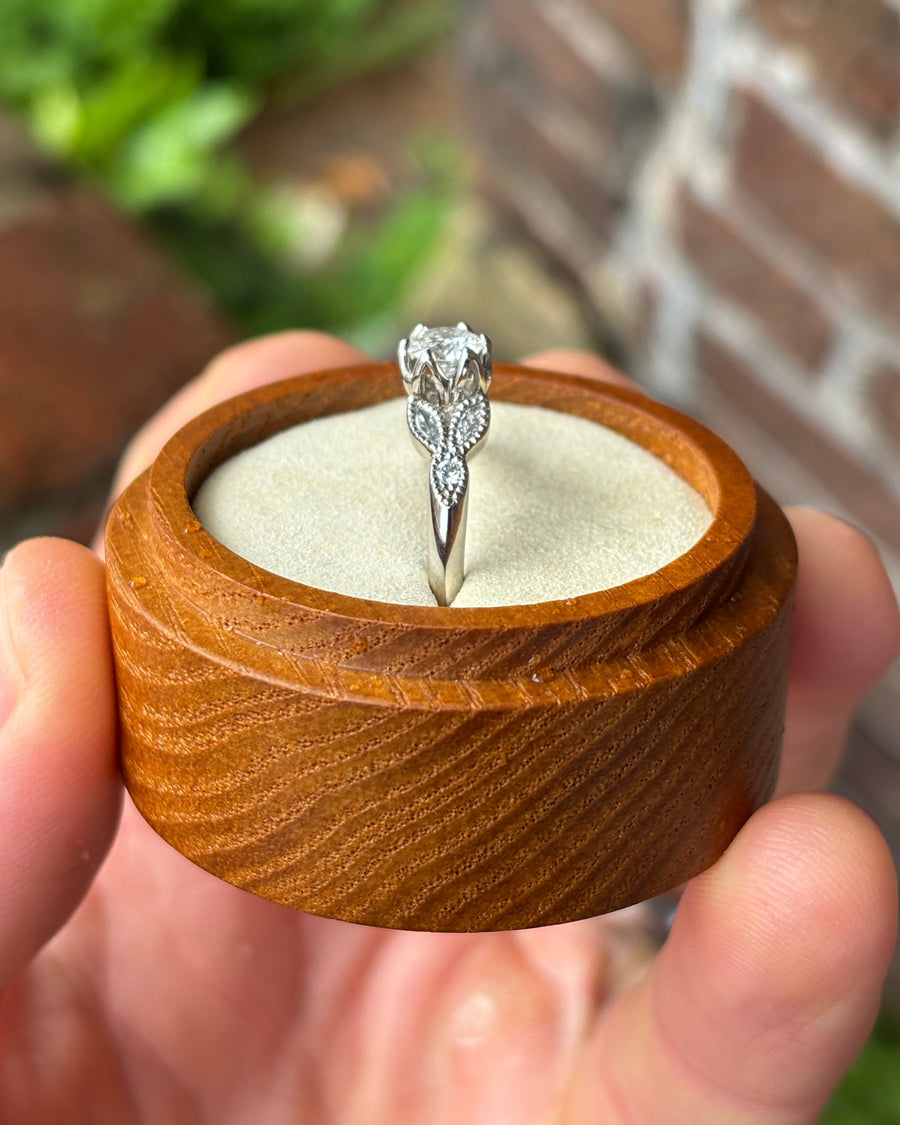 Round cut diamond engagement ring in a silver-toned metal setting with leaf-inspired side details in a wooden box.