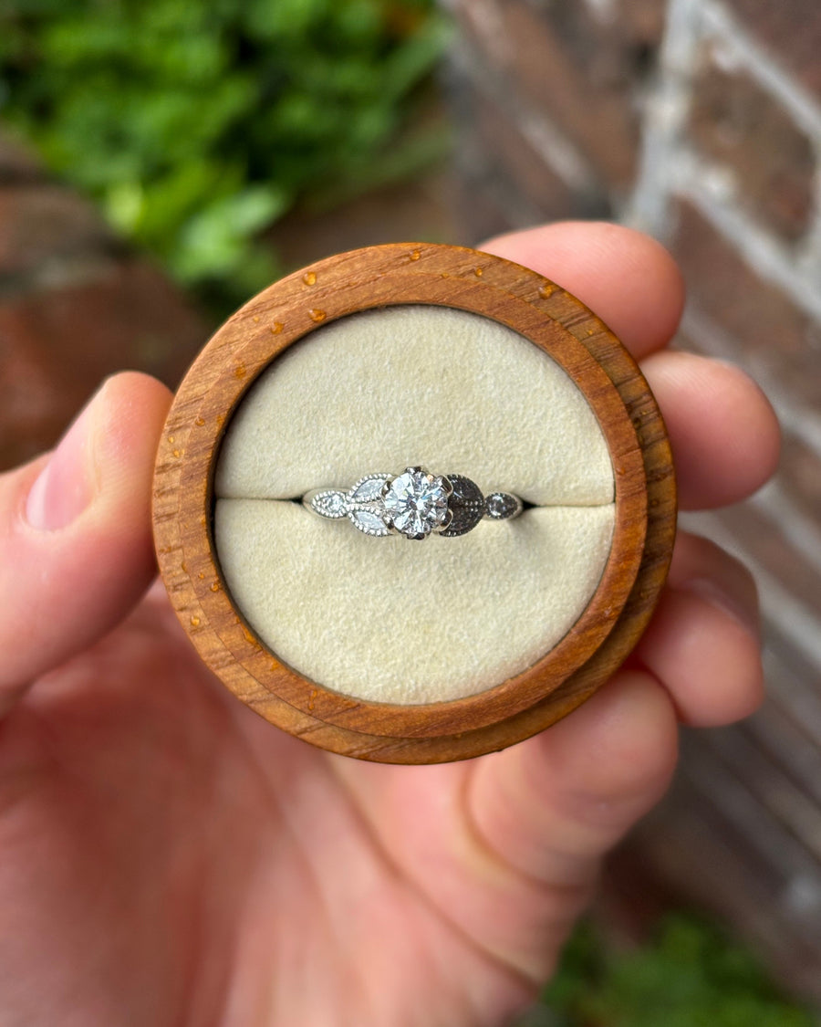 Round cut diamond engagement ring in a platinum vintage setting with leaf-inspired accents in a wooden box.