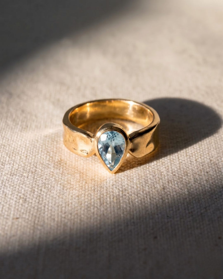 9ct yellow gold side hammered ring with a pear-shaped pale blue aquamarine gemstone.