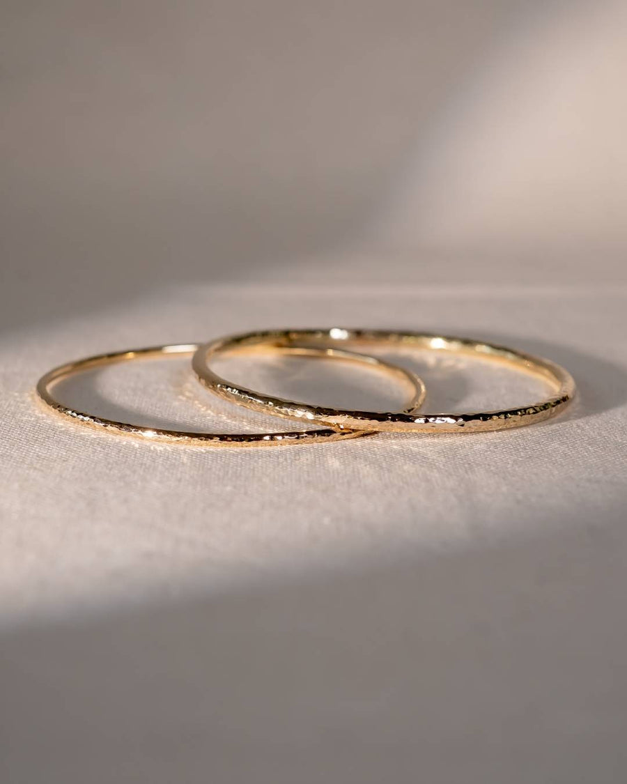 Two handcrafted gold bangles with hammered texture on a soft neutral fabric background
