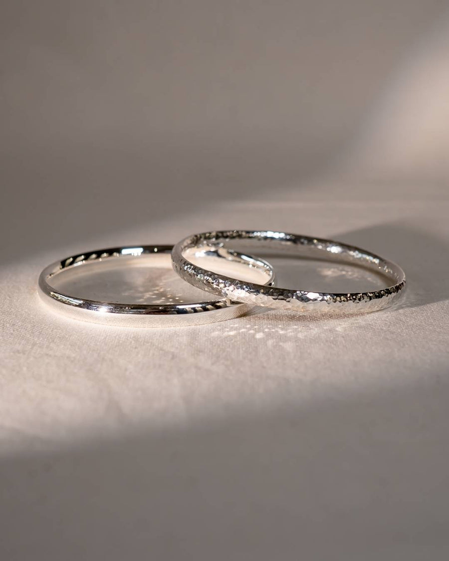 Two handcrafted silver bangles, one with a hammered texture, on a soft fabric surface