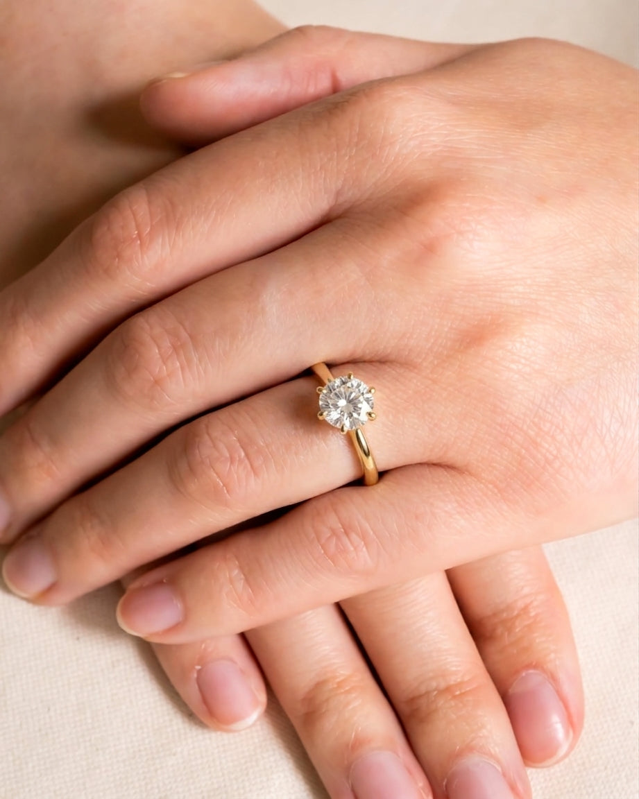 Round cut diamond solitaire engagement ring set in a gold band worn on a finger.