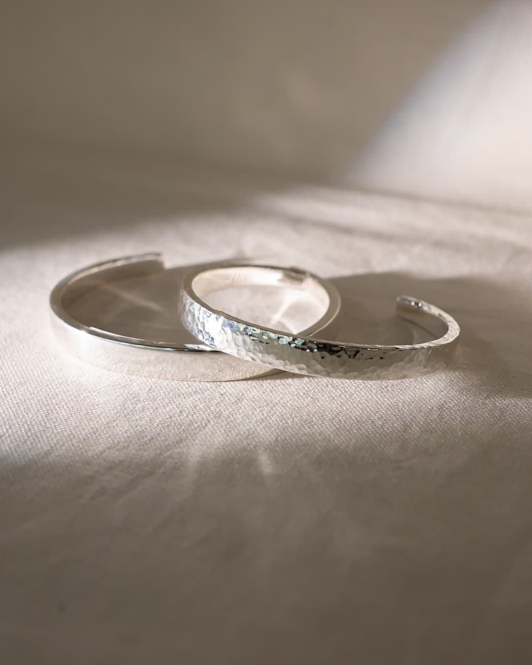 Two handmade silver bangle bracelets, one with a hammered texture, on a light fabric surface.