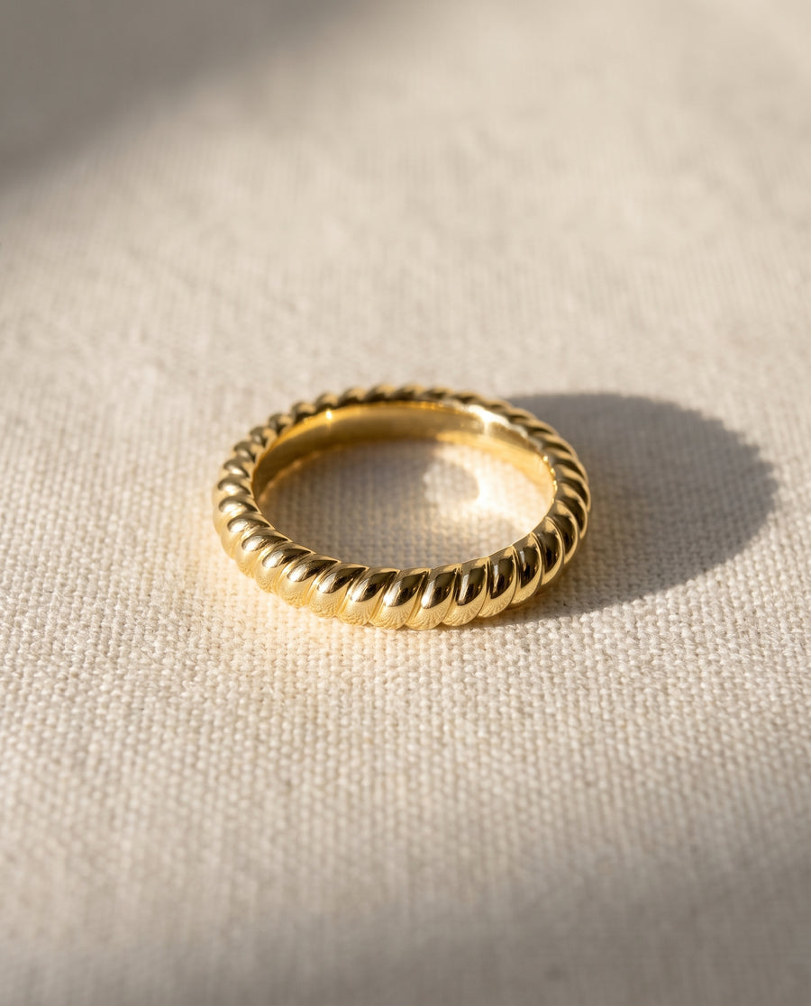 Gold wedding band with a polished, twisted rope design resting on a textured fabric surface.