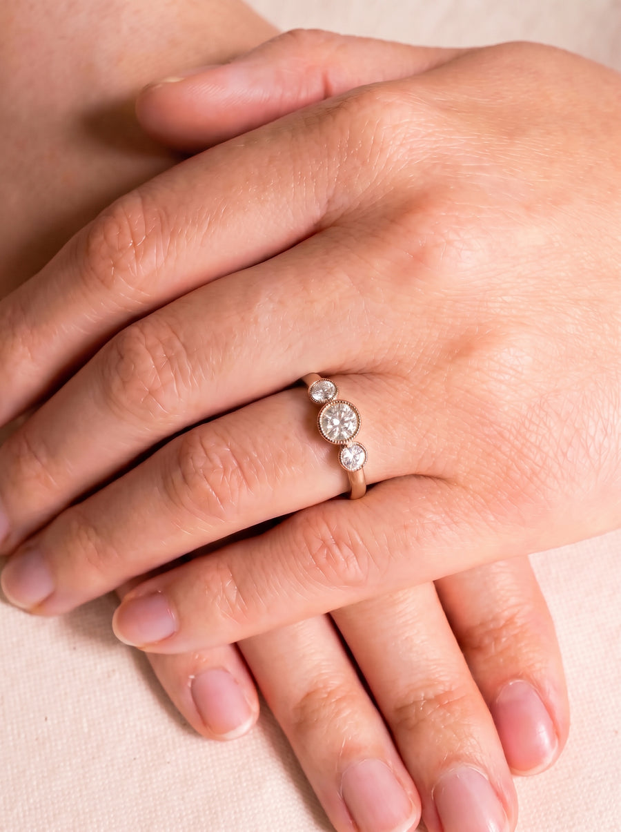 Rose gold trilogy engagement ring featuring three round bezel-set diamonds on a person's finger.