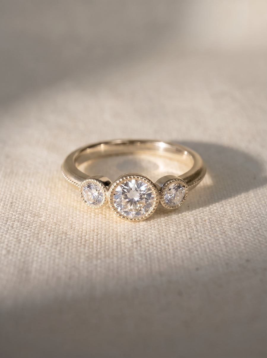 Yellow gold trilogy engagement ring with three round diamonds in milgrain bezel settings on a textured fabric.