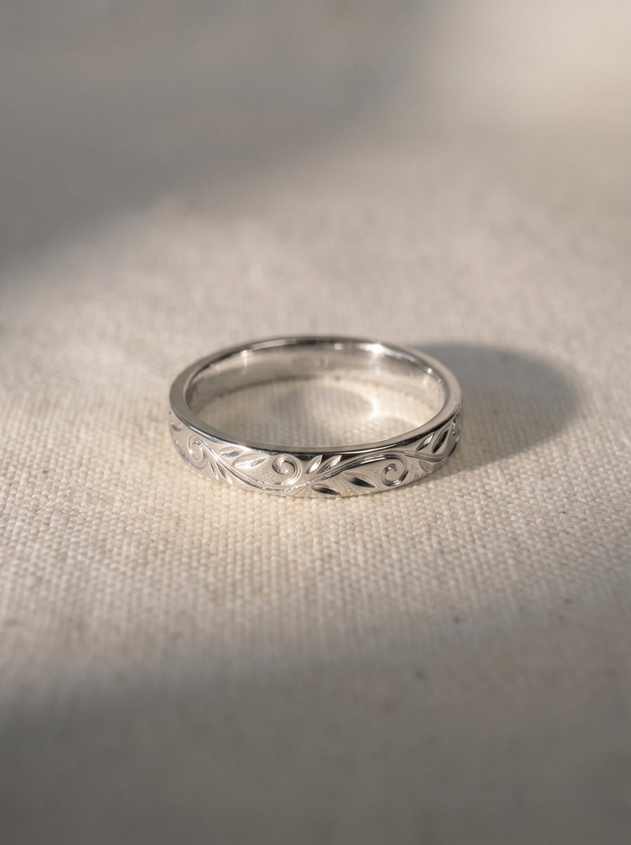 Silver wedding ring with an engraved leaf and scroll pattern on a textured fabric background.