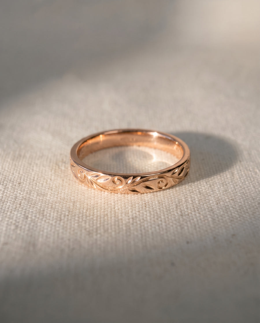 Rose gold wedding ring featuring an intricate engraved leaf and scroll pattern on a textured fabric surface.
