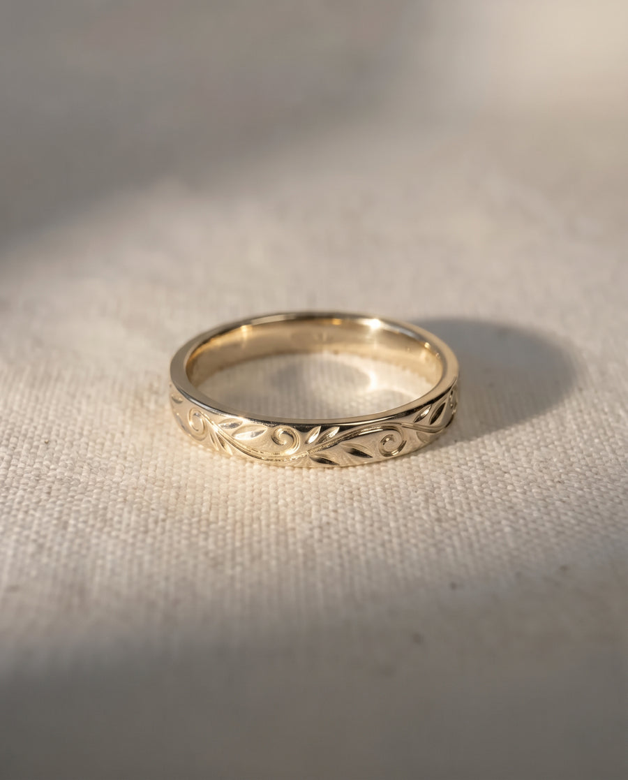 Gold wedding band featuring an engraved floral scroll pattern on a textured fabric background.