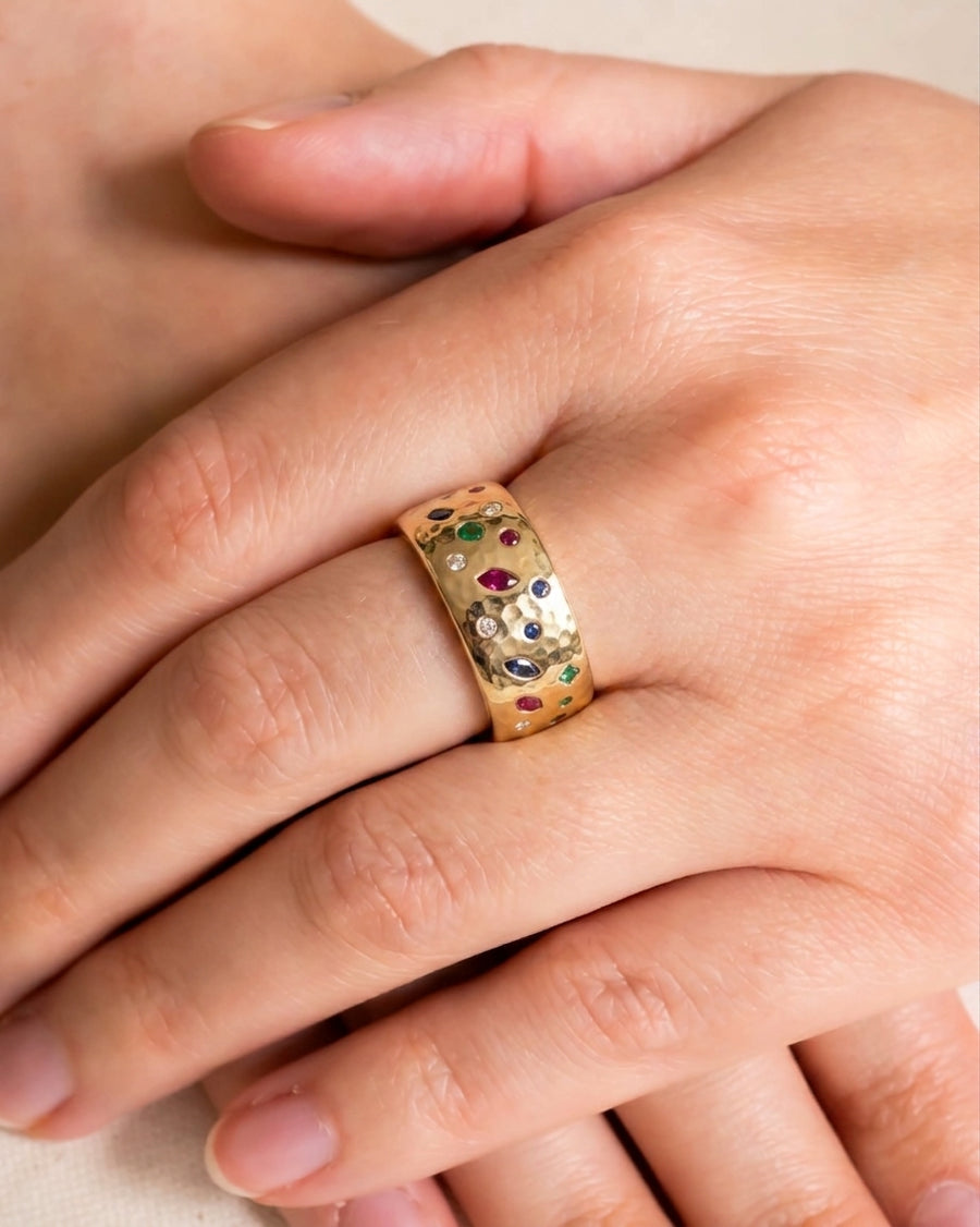 Wide hammered gold ring with scattered sapphire, emerald, ruby, and diamond gemstones.