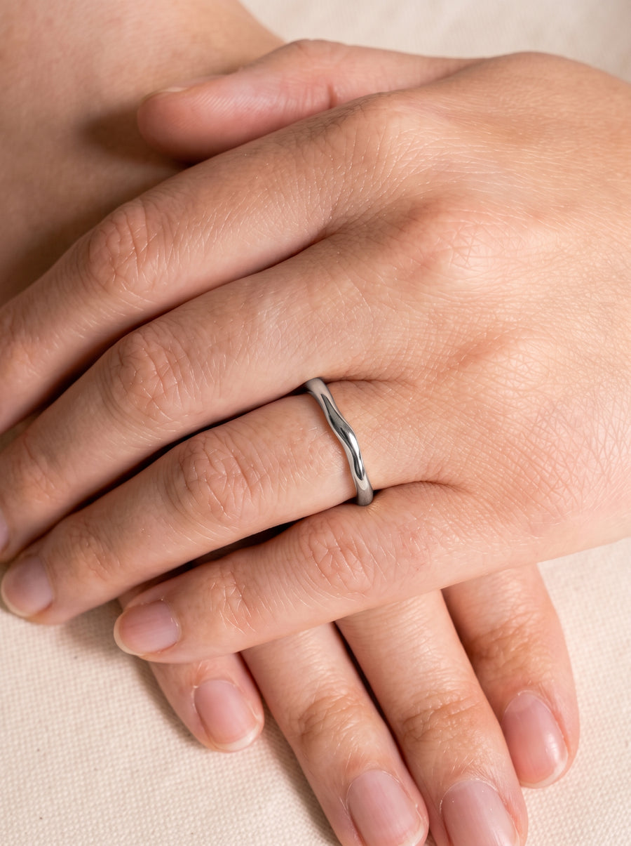 Dainty silver wedding ring with a smooth, uneven wavy band worn on a finger.