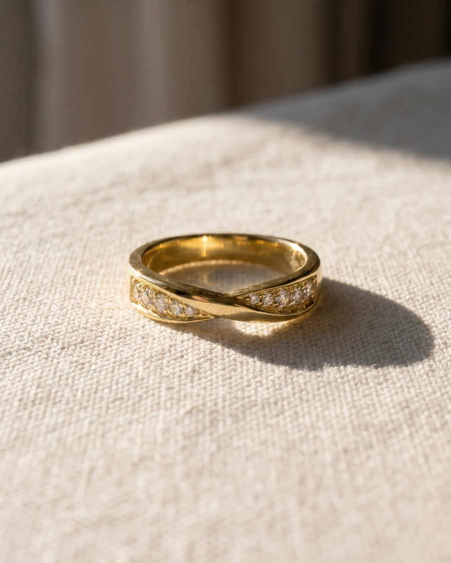 Gold wedding ring with a twisted band design and small embedded diamonds on a textured fabric surface.